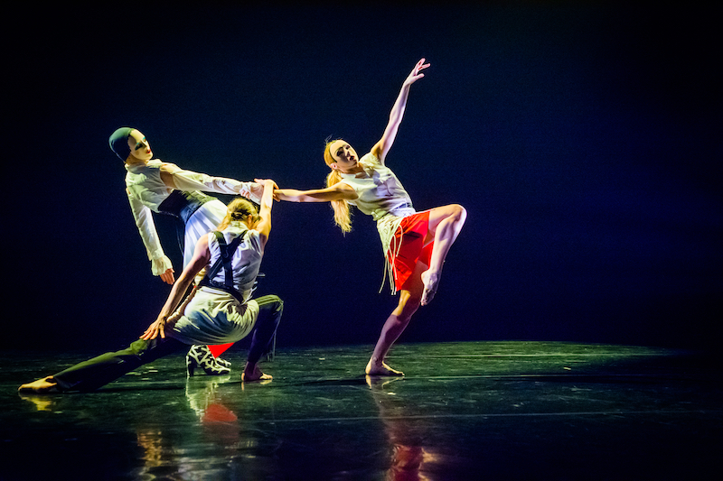 Move and their two dancers wear masks that resemble mimes. They wear white button up shirts with various layers and accessories -- skirts, scarfs, corsets. They hold onto one another by their hands creating a chained effect.